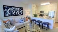 This single-floor residence offers a thoughtfully designed open-concept layout that integrates the living and kitchen areas into one cohesive, functional space. The living area is anchored by a plush white sofa adorned with a mix of patterned and solid accent pillows in navy, white, and coral tones, complemented by a geometric glass-top coffee table that adds a contemporary touch. A bold black-and-white abstract artwork serves as a striking focal point on the main wall, lending the space a modern, curated aesthetic. Warm ambient lighting from a classic table lamp enhances the inviting atmosphere throughout.

The kitchen is positioned adjacent to the living space and features crisp white cabinetry, light-toned countertops, and stainless steel appliances including a built-in microwave and gas range. A raised breakfast bar with sleek navy blue upholstered bar stools provides both a casual dining option and a natural visual boundary between the two zones. A touch of greenery on the counter adds a fresh, lively element to the kitchen. The overall color palette of whites, navy blues, and neutral tones creates a clean, cohesive, and welcoming environment throughout the space.