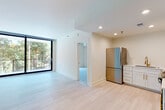 This single-floor unit presents a clean, contemporary design characterized by an all-white palette, light-toned tile flooring, and an abundance of natural light. The open living area is anchored by a large, black-framed floor-to-ceiling sliding glass door that frames lush, tree-lined outdoor views, creating a bright and airy atmosphere throughout the space.

The kitchen is thoughtfully appointed with white shaker cabinetry accented by brushed gold hardware, white quartz countertops, a light tile backsplash, and a suite of stainless steel appliances including a refrigerator, dishwasher, and range. A gold-finish faucet over a stainless steel sink adds a refined touch to the functional layout.

The bedroom features its own floor-to-ceiling black-framed window, offering direct views of surrounding greenery and filling the room with natural light. The bathroom maintains the unit's cohesive aesthetic with gold fixtures, a white vanity with an undermount sink, and light tile flooring. Recessed lighting is present throughout, ensuring consistent, well-distributed illumination in every room. The overall design is modern and minimal, with a cohesive use of materials that creates a polished, move-in-ready environment.