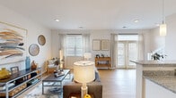 This well-appointed two-floor residence offers a thoughtfully designed layout that balances comfort and functionality. The main living area is open and airy, featuring light hardwood-style flooring, recessed lighting, and an abundance of natural light streaming through multiple windows dressed with sheer curtains and horizontal blinds. The space flows seamlessly from the living room into the kitchen, which is highlighted by granite countertops, a peninsula with pendant lighting, white cabinetry, a double sink, and a built-in dishwasher — all complemented by open wall shelving for added storage and display. The living room itself is generously proportioned, with clean white walls that serve as a neutral backdrop and a layout that accommodates both relaxed lounging and casual entertaining. Bedrooms are carpeted for warmth and comfort, each featuring natural light from window-fitted walls, providing a calm and restful atmosphere. A full bathroom includes a granite-topped vanity with white cabinetry, a single basin sink, and a shower-tub combination. Additional highlights include a spacious walk-in closet with wire shelving on the upper floor, providing ample storage capacity. The dollhouse view reveals a well-organized floor plan with a staircase connecting both levels, making this a versatile and livable space throughout.