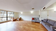 Located at 2700 Saratoga Place in Ottawa, Ontario, this single-floor community space offers a well-organized and functional layout designed for communal use. The open-concept living area is the centrepiece of the space, finished with warm honey-toned hardwood flooring that extends throughout, creating a cohesive and inviting atmosphere. Wall-mounted sconce lighting complements the natural light that pours in through multiple large windows fitted with roller blinds, offering views of the surrounding outdoor greenery and streetscape.

The living area provides generous open floor space, well-suited for gatherings or flexible seating arrangements. A wainscoting detail runs along the lower portion of the walls, adding a classic architectural touch to the otherwise clean, neutral interior. The textured white ceiling and soft cream-toned walls contribute to a bright, airy feel.

The kitchen is clearly defined and efficiently laid out, featuring warm wood cabinetry, dark granite-style countertops, a stainless steel sink, and white appliances including a refrigerator, stove, and microwave. Light ceramic tile flooring grounds the kitchen area, providing a practical contrast to the hardwood in the main room. A pass-through counter connects the kitchen to the living space, supporting easy service and interaction during events. Overall, the space is clean, functional, and well-maintained.