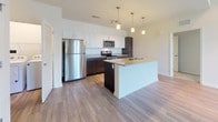 This well-designed single-floor residence offers a thoughtful layout that balances open communal spaces with comfortable private quarters. The heart of the home is an open-concept kitchen and living area, where warm hardwood-style flooring flows throughout. The kitchen is outfitted with a full suite of stainless steel appliances, including a refrigerator, range, and microwave, complemented by a two-tone cabinet design featuring white upper cabinets and dark lower cabinets. Granite countertops extend across the workspace and onto a central island, which is illuminated by a trio of pendant lights hanging overhead — making it an ideal spot for casual dining or meal prep. The open living area benefits from sliding glass door access and a ceiling fan, contributing to a bright and airy atmosphere. The bedrooms are carpeted for comfort, each equipped with a ceiling fan and a window dressed with blinds that allow for natural light. A hallway connects the private spaces to the rest of the unit. The bathrooms are finished with wood-tone vanities, granite countertops, and tub-shower combinations. A separate laundry area with washer and dryer hookups adds practical convenience to this well-rounded residence.