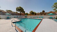This outdoor community pool area offers a welcoming and functional recreational space designed for relaxation and leisure. At its center is a large, rectangular swimming pool with crystal-clear turquoise water, framed by classic brick-red tile edging that adds a distinctive architectural detail. The pool is equipped with stainless steel entry rails and lane markers, making it accessible and practical for swimmers of all levels.

Surrounding the pool is a broad, poured concrete sun deck that provides ample space for lounging. Numerous teal-colored chaise lounge chairs and seating sets are arranged around the perimeter, complemented by several teal market umbrellas that offer shaded areas for those seeking relief from the sun. The deck's generous proportions allow for comfortable movement and social gathering throughout the space.

The entire area is enclosed by a white wooden privacy fence, creating a defined and secure environment. Mature trees visible beyond the fence provide a natural backdrop and contribute to a pleasant, park-like ambiance. On one side of the pool, an adjacent single-story building with a pitched shingle roof and large windows appears to serve as a clubhouse or amenity facility, offering convenient access to additional services. The open sky above and surrounding greenery complete the inviting outdoor atmosphere of this community amenity.