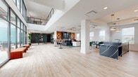 Located at 9350 District Way in Mason, Ohio, this single-floor amenity space offers a generously sized, thoughtfully designed environment that seamlessly blends social and wellness functions. The open-plan lounge area commands attention with soaring double-height ceilings, light hardwood flooring, and an upper mezzanine level visible overhead. Large floor-to-ceiling windows flood the interior with natural light and provide views of the surrounding district. The lounge is appointed with an eclectic mix of emerald green seating, a bar-height counter with pendant lighting, and a media wall framed by warm, earth-toned accent walls — creating distinct social zones within the expansive layout. A decorative ceiling installation featuring sculptural bird motifs adds an artistic focal point, while bold abstract artwork and lush indoor plants contribute to the vibrant, curated atmosphere. A beverage and refreshment station is conveniently integrated near a glass wall leading to an outdoor terrace. The dedicated exercise room features a teal-accented ceiling, patterned carpet flooring, ceiling fans, and an extensive lineup of cardio equipment including treadmills, ellipticals, stationary bikes, and stair climbers. Natural light pours in through a row of windows along the gym wall, creating an energizing workout environment. Together, these spaces offer a polished, amenity-rich experience.