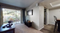 This single-story residence offers a practical and well-organized layout across several distinct living spaces. The living room serves as the central gathering area, finished with plush carpet and neutral-toned walls. A wall-mounted television anchors the room, while a sliding glass door with teal curtain panels fills the space with natural light and provides direct access to a private outdoor patio, offering views of a fenced yard and surrounding greenery.

Adjacent to the living room, a dedicated dining area features wood-look flooring and a tray ceiling detail with overhead lighting, creating a defined yet open feel. The kitchen is efficiently arranged in a U-shaped configuration, showcasing dark cabinetry with brushed hardware, light countertops, wood-look flooring, and black appliances including a dishwasher and range, along with a stainless steel refrigerator. A window above the counter brings in additional natural light.

The two bedrooms are carpeted and neutrally finished, each offering comfortable proportions. One bedroom includes a window with blinds and curtain panels. The bathrooms feature wood-look flooring, dark vanity cabinetry with light countertops, and a tub-shower combination. One bathroom also includes open shelving and a louvered-door storage closet, adding functional storage to the space.