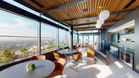 This sophisticated rooftop amenity space is defined by its striking architectural design and panoramic city views. Floor-to-ceiling glass walls wrap around the primary lounge area, flooding the interior with abundant natural light and framing expansive views of the surrounding urban skyline. A warm slatted wood ceiling adds texture and visual depth, contrasting beautifully with the dark structural steel framing and light tile flooring below.

The main lounge area is furnished with round tables and modern terracotta-toned chairs arranged to maximize both social interaction and view enjoyment. Oversized globe pendant lights hang from the ceiling, providing warm ambient illumination that complements the natural light. Access to an outdoor terrace extends the usable space, further enhancing the connection to the cityscape.

A well-appointed kitchenette serves as a functional centerpiece, featuring sage green vertical tile work, dark cabinetry, open shelving, a sink, and under-counter refrigeration units. A solid wood bar-height counter with ornate metal bar stools provides an inviting spot for casual dining or socializing.

Tucked away near the entrance is a cozy booth seating nook, upholstered in rich brown leather and set against a green tile accent wall with warm LED underlighting, offering a more intimate setting within the broader space. The overall ambiance is polished, modern, and welcoming.