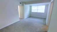 This single-floor residential space presents a clean, move-in-ready environment defined by its neutral color palette and practical design. The main living area is generously proportioned, featuring wall-to-wall plush carpeting in a warm beige tone that adds comfort underfoot and a sense of coziness to the space. Soft, light-green walls contribute to a calm and airy ambiance throughout.

A large window in the main room allows an abundance of natural light to fill the interior, brightening the space and offering a view of the surrounding outdoor environment. The ceiling-mounted HVAC vent ensures efficient climate control, while clean white baseboards and smooth walls reflect a well-maintained interior.

An open doorway visible from the main living area leads to an adjoining room or hallway, suggesting a functional and connected floor plan that allows for easy movement between spaces. The transition area features light-colored flooring, contrasting with the carpeted living zone and providing a practical surface for higher-traffic areas.

Overall, this space offers a straightforward, comfortable layout with a bright and neutral aesthetic, making it highly adaptable for a variety of furnishing styles and personal preferences.
