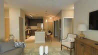 This single-floor residence showcases a thoughtfully designed open-concept layout that integrates the kitchen, dining, and living areas into one cohesive space. The kitchen is a standout feature, equipped with dark espresso cabinetry, stainless steel appliances, and light stone countertops that contrast beautifully with the rich cabinet tones. A central kitchen island doubles as a breakfast bar with barstool seating, making it ideal for casual dining or entertaining. Pendant lighting above the island adds a warm, inviting glow that complements the recessed and track lighting throughout. Light-toned ceramic tile flooring spans the open areas, reflecting the warm ambient lighting and contributing to a bright, welcoming atmosphere. The living area transitions naturally from the kitchen, featuring a decorative area rug with a coastal-inspired pattern and elegant accent seating. A spacious walk-in closet is visible from the main hallway, offering generous storage. The overall ambiance is modern yet comfortable, with warm neutral tones, clean lines, and a well-balanced mix of functional and decorative elements throughout.