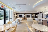 Located at 101 E Camino Real in Boca Raton, Florida, this upscale amenity lounge delivers a refined, contemporary atmosphere across a well-appointed single-floor layout. The open-plan design seamlessly integrates multiple seating zones, creating a versatile space suited for both social gatherings and relaxed lounging.

The lounge area is anchored by a plush cream sectional sofa and complemented by multiple round dining tables paired with sculptural cream and tan chairs, offering generous seating throughout. Large floor-to-ceiling windows and glass entry doors flood the interior with natural light while providing direct views of the outdoor patio and pool area beyond.

A standout architectural feature is the circular recessed ceiling detail in the kitchenette zone, beneath which a dramatic sputnik-style chandelier adds a bold focal point. The kitchenette itself features a curved white quartz island with modern bar stools, warm wood cabinetry, stainless steel appliances, and a herringbone-tiled backsplash. Ambient cove lighting runs along the ceilings throughout, casting a warm golden glow that enhances the sophisticated atmosphere.

Dark wood wall paneling, a wall-mounted flat-screen TV, built-in shelving, and carefully curated abstract artwork contribute to the polished, hotel-inspired aesthetic. The flooring transitions between large-format marble-effect tile and light wood-tone carpeting, further defining each functional zone within the space.