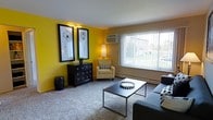 This single-floor space showcases a thoughtfully designed living room that immediately draws attention with its bold yellow accent wall, creating a warm and energetic focal point. Two framed botanical art pieces mounted on the yellow wall add a decorative touch, complementing the room's overall aesthetic. The living area is carpeted throughout, contributing to a comfortable and inviting atmosphere.

A large window with vertical blinds dominates one wall, allowing an abundance of natural light to fill the room while offering views of the surrounding outdoor area. A wall-mounted air conditioning unit is conveniently positioned nearby, ensuring year-round comfort. The neutral-toned walls surrounding the accent wall provide a balanced contrast, keeping the space feeling open and airy.

The room comfortably accommodates a seating arrangement that includes a sofa and an accent chair with striped cushions, centered around a sleek, modern coffee table. A dark dresser with circular hardware serves as both storage and a display surface along the accent wall. A baseboard heating unit runs along the exterior wall, further emphasizing the space's practical and functional design.