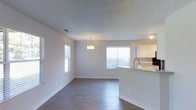 This renovated two-bedroom, two-bathroom single-floor residence offers a thoughtfully designed open-concept layout that seamlessly connects the living, dining, and kitchen areas. The living space features durable wood-style flooring throughout, contributing to a clean, contemporary aesthetic. Multiple windows fitted with blinds allow generous natural light to fill the interior, creating a bright and welcoming atmosphere.

The kitchen is positioned adjacent to the main living area and is separated by a peninsula-style counter with an integrated sink, providing both a functional workspace and a casual dining or entertaining surface. White cabinetry above offers ample storage, while a pendant light fixture adds a stylish focal point above the counter. The textured ceiling adds subtle dimension to the space.

The open flow between the kitchen and living area maximizes the sense of space, making the layout feel practical and versatile. The neutral color palette of whites and soft grays throughout the unit provides a clean backdrop that complements a variety of décor styles. Overall, this renovated unit combines modern updates with functional design across a well-proportioned, single-level floor plan.
