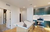 Ruby Slipper is a thoughtfully designed 1-bedroom, 1-bathroom residence offering 640 square feet of contemporary living space across a single floor. The open-plan kitchen and living area serves as the heart of the unit, showcasing bold emerald green cabinetry with under-cabinet LED lighting, a dramatic marble-veined island countertop, a farmhouse-style stainless steel sink, a herringbone tile backsplash, and stainless steel appliances. Light hardwood flooring flows throughout the main living areas, enhancing the bright, airy atmosphere created by large black-framed windows. A glass-paneled door opens to a private balcony, offering elevated views of the surrounding cityscape and neighborhood. The 120-square-foot bedroom features plush carpet, a ceiling fan, and a generously sized walk-in closet with wire shelving on multiple walls. The bathroom is a design standout, featuring bold black-and-white geometric tile flooring, a sleek wood-toned vanity with brass hardware and a white quartz countertop, black matte fixtures, and a glass-enclosed shower adorned with deep green stacked tile. Recessed lighting throughout the unit completes the polished, modern aesthetic.