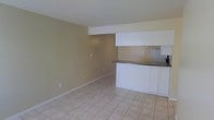 This single-floor space features an open-concept layout that seamlessly connects the living area to the kitchen. The living space is finished with light-toned ceramic tile flooring that runs throughout, contributing to a clean and cohesive aesthetic. Neutral beige walls provide a versatile backdrop, while white baseboards offer a crisp contrast.

The kitchen is defined by a raised breakfast bar with a dark countertop, supported by white cabinetry beneath. A soffit overhead frames the kitchen space, giving it a structured, intentional boundary within the open floor plan. Stainless steel appliances, including a refrigerator, are visible in the background, adding a modern touch. A sink with basic fixtures completes the functional kitchen setup.

The textured popcorn ceiling is consistent throughout the visible areas, and recessed structural elements add subtle architectural definition. The overall ambiance is practical and straightforward, with a simple, functional design that offers an easy-to-maintain living environment. The open layout allows for flexible furniture arrangement and makes the space feel airy and uncluttered.