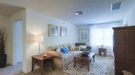 This single-floor space showcases a thoughtfully designed living room that serves as its central feature. The room is finished with soft, neutral white walls that create a clean and airy atmosphere, complemented by light-colored carpeting underfoot. A large window dressed with sheer white curtains allows generous natural light to fill the space, enhancing its open and welcoming feel.

The living area is arranged around a comfortable light beige sofa accented with decorative pillows in blue and floral patterns, paired with a classic upholstered armchair in a deeper tone. A wooden coffee table with a lattice-style base anchors the seating area, sitting atop a patterned area rug that adds visual interest to the floor. Matching end tables with similar lattice detailing provide functional surface space, one of which holds a stylish table lamp that contributes warm ambient lighting.

Wall décor includes two framed prints displayed side by side, alongside a botanical-themed framed piece near the window, adding character without overwhelming the room. A ceiling vent is centrally placed, indicating a climate control system. Overall, the space projects a calm, comfortable, and well-organized residential environment.