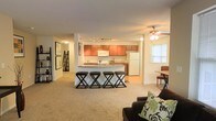 This single-floor space features an open-concept layout that seamlessly connects the living, dining, and kitchen areas. The kitchen is well-appointed with warm medium-toned wood cabinetry, a white refrigerator, and a microwave, complemented by track lighting overhead. A peninsula-style breakfast bar with seating for three on sleek black stools serves as a natural divider between the kitchen and the living area, making it ideal for casual dining or entertaining. Adjacent to the kitchen, a small dining nook with a table and chairs sits beside a window, allowing natural light to filter in. The living area features plush wall-to-wall carpeting and is anchored by a dark sofa accented with decorative pillows. A ceiling fan with a light fixture provides both comfort and illumination. A lean-to bookshelf and framed wall art add character near the entryway corridor, which leads further into the space. The overall layout is practical and cohesive, with a neutral color palette throughout that creates a comfortable and inviting atmosphere suitable for everyday living.