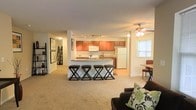 This single-floor space features an open-concept layout that seamlessly connects the living, dining, and kitchen areas. The kitchen is well-appointed with warm medium-toned wood cabinetry, a white refrigerator, and a microwave, complemented by track lighting overhead. A peninsula-style breakfast bar with seating for three on sleek black stools serves as a natural divider between the kitchen and the living area, making it ideal for casual dining or entertaining. Adjacent to the kitchen, a small dining nook with a table and chairs sits beside a window, allowing natural light to filter in. The living area features plush wall-to-wall carpeting and is anchored by a dark sofa accented with decorative pillows. A ceiling fan with a light fixture provides both comfort and illumination. A lean-to bookshelf and framed wall art add character near the entryway corridor, which leads further into the space. The overall layout is practical and cohesive, with a neutral color palette throughout that creates a comfortable and inviting atmosphere suitable for everyday living.