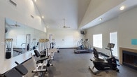 This fitness center offers a spacious, single-floor workout environment designed to accommodate a range of exercise routines. The room features notably high vaulted ceilings that lend an open, airy feel to the space, preventing any sense of confinement during workouts. Recessed lighting and a ceiling fan ensure the area remains well-lit and ventilated throughout use. A large wall-mounted mirror along one side visually expands the room while allowing users to monitor their form during exercise. The flooring consists of durable, dark rubberized material well-suited for fitness activity. Strength training equipment, including a multi-station cable machine and various weight benches, occupies one section of the floor. Cardio machines, such as a stair climber and a recumbent bike, are positioned throughout the remaining space, offering diverse workout options. A wall-mounted television provides entertainment during exercise sessions. Natural light filters in through a window on one side of the room, contributing to the bright and motivating atmosphere. The neutral color palette of the walls and ceiling keeps the environment clean and focused.