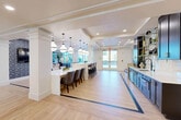 Located at Presidential Way in West Palm Beach, FL, this generously sized single-floor amenity space offers a refined, coastal-contemporary environment spanning a substantial footprint. The open-concept layout flows seamlessly between distinct functional zones, unified by light-toned hardwood-style flooring accented with contrasting dark inlay borders.

A central highlight is the elongated white quartz island bar, illuminated by a dramatic row of teardrop pendant lights and flanked by stylish counter-height seating. Adjacent to the island, a full wet bar features deep navy cabinetry with brass hardware, a marble backsplash, open glass-and-gold shelving, and an undermount sink — providing an elevated hospitality experience.

The lounge area accommodates comfortable seating arrangements beneath coffered ceilings with recessed lighting and a statement circular chandelier. Navy built-in display cabinetry and a mounted flat-screen television anchor the space visually. White structural columns define zones without disrupting the open flow.

Floor-to-ceiling windows and glass-paneled double doors flood the interior with natural light and provide views of the surrounding outdoor landscape. The overall ambiance is polished yet approachable, combining modern design elements with warm, inviting finishes suited to the South Florida setting.