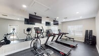 This single-floor fitness center in Mobile, Alabama offers a comprehensive workout environment designed to accommodate a variety of exercise routines. The space is organized into distinct functional zones, each thoughtfully arranged to maximize usability and comfort.

The primary cardio area features multiple treadmills and elliptical machines from Star Trac and similar brands, all positioned facing ceiling-mounted flat-screen televisions for an engaging workout experience. Large wall-to-wall mirrors line one side of the room, visually expanding the space and allowing users to monitor their form during exercise. The flooring throughout consists of durable dark rubber gym matting, providing traction and impact absorption.

A dedicated cycling area houses several Schwinn stationary bikes, positioned apart from the cardio machines to allow ample space for movement. Natural light filters in through multiple windows fitted with horizontal blinds, contributing to a bright and welcoming atmosphere, while recessed ceiling lighting ensures consistent illumination throughout.

A separate free weights alcove provides a quieter strength-training environment, equipped with a full dumbbell rack ranging across multiple weight increments and an adjustable workout bench. White walls and neutral tones throughout the facility create a clean, modern aesthetic that keeps the focus on fitness.