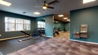 This single-level fitness center is thoughtfully designed to accommodate a variety of workout disciplines across several distinct functional zones. The space is unified by a consistent design palette of deep teal walls, white baseboards, and a geometric-patterned tile floor in muted earth tones, creating a modern and energizing atmosphere throughout.

The main open area provides ample room for free weight training, featuring a tiered dumbbell rack, a weight bench, and a cable machine. Rubber flooring mats define the strength training zone, offering both safety and durability. Large wall mirrors line multiple sections of the facility, visually expanding the space and allowing users to monitor their form.

A dedicated cardio room houses treadmills, ellipticals, and a stair climber, accompanied by a wall-mounted flat-screen television for entertainment during workouts. An adjacent cycling area contains a row of stationary spin bikes positioned in front of floor-to-ceiling mirrors. Rowing machines occupy a quieter corner of the facility, ideal for low-impact full-body conditioning.

Overhead, recessed panel lighting and ceiling fans provide consistent illumination and air circulation. A built-in water fountain station is conveniently accessible near the main corridor. Natural light filters in through windows, and a glass-paneled door offers access to an exterior area, contributing to the facility's open and well-ventilated feel.