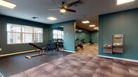 This single-level fitness center is thoughtfully designed to accommodate a variety of workout disciplines across several distinct functional zones. The space is unified by a consistent design palette of deep teal walls, white baseboards, and a geometric-patterned tile floor in muted earth tones, creating a modern and energizing atmosphere throughout.

The main open area provides ample room for free weight training, featuring a tiered dumbbell rack, a weight bench, and a cable machine. Rubber flooring mats define the strength training zone, offering both safety and durability. Large wall mirrors line multiple sections of the facility, visually expanding the space and allowing users to monitor their form.

A dedicated cardio room houses treadmills, ellipticals, and a stair climber, accompanied by a wall-mounted flat-screen television for entertainment during workouts. An adjacent cycling area contains a row of stationary spin bikes positioned in front of floor-to-ceiling mirrors. Rowing machines occupy a quieter corner of the facility, ideal for low-impact full-body conditioning.

Overhead, recessed panel lighting and ceiling fans provide consistent illumination and air circulation. A built-in water fountain station is conveniently accessible near the main corridor. Natural light filters in through windows, and a glass-paneled door offers access to an exterior area, contributing to the facility's open and well-ventilated feel.