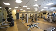 Located at 400 S Military Hwy in Virginia Beach, VA, this expansive fitness center offers a well-organized, single-floor layout designed to accommodate a full range of workout needs. The facility is finished with durable, dual-tone rubberized flooring in tan and gray — ideal for high-traffic athletic use — and features a suspended drop-tile ceiling with recessed panel lighting that ensures bright, even illumination throughout the space.

The cardio zone is generously equipped with multiple treadmills, elliptical trainers, stationary bikes, and stair climbers, several of which are positioned along windowed walls fitted with slatted blinds, allowing natural light to fill the area. A wall-mounted flat-screen television is positioned for visibility across the floor, enhancing the workout experience.

The strength training area is equally well-appointed, featuring a variety of Precor selectorized cable and resistance machines, free-weight benches, dumbbells, and kettlebells arranged on dedicated rack stations. Full-length mirrors line key sections of the walls, creating a sense of openness and allowing users to monitor their form.

The layout provides ample open floor space between equipment stations, supporting safe and comfortable movement throughout. The neutral color palette, clean lines, and organized arrangement contribute to a professional and functional fitness environment suitable for users of all fitness levels.