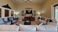 This single-floor clubhouse-style facility is designed to offer a refined and comfortable communal experience. The main lounge area is a standout space, featuring soaring tray ceilings adorned with crown molding and a chandelier, flanked by decorative columns that define the room's layout. Neutral warm-toned walls, tile flooring, and a patterned area rug anchor the seating arrangement, which centers around a large flat-screen television mounted above a rich wood media cabinet. Plush sofas, upholstered accent chairs, and coordinated décor contribute to an inviting atmosphere. The kitchen is well-equipped with abundant warm wood cabinetry, dark granite-style countertops, full-size appliances including a refrigerator and range, a peninsula with bar seating, and pendant lighting overhead. A game room accommodates a full-size billiards table beneath ample ceiling height, with arched entry doors allowing natural light to filter in. A hallway features decorative wall sconces, an artistic mirror gallery arrangement, and a mounted water fountain, offering functional amenities with thoughtful design details throughout the space.