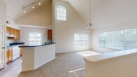 This single-floor residence makes a striking first impression with its dramatic vaulted ceilings that soar above the open-concept living area, creating an expansive and airy atmosphere. Track lighting and a ceiling fan complement the abundance of natural light streaming in through multiple large windows fitted with blinds, including a prominent arched window positioned high on the vaulted wall. The living space flows seamlessly into a kitchen featuring warm wood cabinetry, dark countertops, a breakfast bar peninsula, and white appliances. The kitchen benefits from its own window and tile flooring, offering a practical and functional workspace. Carpeted bedrooms are generously sized, each offering ceiling fans and wide windows that frame outdoor greenery, filling the rooms with soft, natural light. The bathrooms are finished with tile flooring, wood vanity cabinetry topped with granite-style countertops, and a tub-shower combination with white tile surround. Walk-in closets with built-in shelving and hanging rods provide ample storage adjacent to the bathrooms. A staircase with a white-painted banister adds architectural interest to the upper portion of the main living area. Throughout, neutral tones and clean lines create a bright, welcoming ambiance.