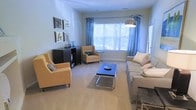This single-floor living space centers around a spacious, light-filled living room designed for both comfort and style. The focal point is a wide bay window dressed with flowing blue curtains, allowing generous natural light to fill the room while offering views of surrounding greenery outdoors. The neutral carpeted flooring provides a warm, cohesive base throughout the space. Seating is thoughtfully arranged, featuring a large, cushioned sofa adorned with accent pillows in soft tones, complemented by two tan upholstered armchairs that add warmth and visual balance. A sleek, modern coffee table with a mirrored or glass surface and chrome legs anchors the seating area, adding a contemporary touch. A dark wood shelving unit along one wall provides functional storage and display space, topped with decorative accessories. A gallery-style arrangement of framed artwork above the unit adds a curated, personalized feel to the room. An arc floor lamp with a metallic shade adds ambient lighting and a stylish vertical element. Overall, the space conveys a bright, welcoming ambiance with a balanced blend of modern and transitional design elements.