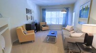This single-floor living space centers around a spacious, light-filled living room designed for both comfort and style. The focal point is a wide bay window dressed with flowing blue curtains, allowing generous natural light to fill the room while offering views of surrounding greenery outdoors. The neutral carpeted flooring provides a warm, cohesive base throughout the space. Seating is thoughtfully arranged, featuring a large, cushioned sofa adorned with accent pillows in soft tones, complemented by two tan upholstered armchairs that add warmth and visual balance. A sleek, modern coffee table with a mirrored or glass surface and chrome legs anchors the seating area, adding a contemporary touch. A dark wood shelving unit along one wall provides functional storage and display space, topped with decorative accessories. A gallery-style arrangement of framed artwork above the unit adds a curated, personalized feel to the room. An arc floor lamp with a metallic shade adds ambient lighting and a stylish vertical element. Overall, the space conveys a bright, welcoming ambiance with a balanced blend of modern and transitional design elements.