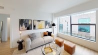 This contemporary single-floor residence offers a thoughtfully designed open layout with clean lines, white walls, and warm wood-tone flooring throughout. The living area is bathed in natural light through expansive floor-to-ceiling windows that frame urban cityscape views, and is complemented by a wall-mounted television and modern furnishings. The open-concept kitchen features gray cabinetry, a white quartz countertop peninsula with bar seating, pendant lighting, a tile backsplash, and a full suite of stainless steel appliances including a gas range, built-in microwave, and refrigerator. The primary bedroom is a serene retreat with direct access to a walk-in closet and an en-suite bathroom featuring a glass-enclosed walk-in shower, white tile surround, and a clean white vanity. The second bedroom benefits from a large window with city views and warm wood flooring, while the second bathroom includes a soaking tub, white tile surround, and a bright white vanity with a large mirror. Throughout the residence, the design maintains a cohesive modern aesthetic with neutral tones, curated artwork, and an overall bright, airy ambiance.