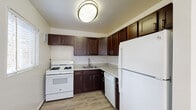 This single-floor unit offers a well-organized layout with distinct living, sleeping, and kitchen zones. The kitchen is equipped with rich dark-brown shaker-style cabinetry extending to the upper walls, light granite-look countertops, and wood-look vinyl flooring. White appliances include a gas range, dishwasher, and full-size refrigerator, complemented by a stainless steel sink and flush ceiling light fixture.

The living areas feature neutral carpeting and crisp white walls, creating a bright and open atmosphere. Multiple windows fitted with horizontal blinds allow natural light to fill the space, and a baseboard heating unit runs beneath the main window. A transitional area with wood-look flooring connects the kitchen to the broader living space, defined by a partial wall partition.

The bedroom is generously sized with plush carpeting, a ceiling fan with integrated lighting, and a window overlooking greenery. Two side-by-side mirrored sliding-door closets provide ample storage.

The bathroom is a standout feature, showcasing floor-to-ceiling marble-patterned tile surrounding a full soaking tub, a small window for natural light, and clean white fixtures that complete the polished aesthetic.