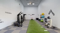 This fitness center is a comprehensively outfitted facility designed to support a wide range of training styles and fitness goals. The space spans two connected areas, unified by a checkered gray and blue tile flooring that lends a clean, athletic aesthetic throughout.

The primary training room features soaring ceilings and bright overhead lighting, creating an open and energizing environment. A central artificial turf lane runs the length of the room, marked with yardage lines for sprint and agility drills. Flanking the turf are a multi-functional cable machine and a heavy tire, ideal for strength and functional training. Colorful medicine balls are neatly stored on a wall-mounted rack, alongside foam rollers, resistance bands, and exercise mats. Plyometric boxes of varying heights are stationed in the corner, and a weight plate rack provides additional resistance training options. Motivational phrases such as "No Pain No Gain," "Strength & Growth," and "Be Stronger Than Your Excuses" are displayed prominently on the crisp white walls.

The adjacent cardio room connects seamlessly through an arched passageway and offers treadmills, a rowing machine, an adjustable weight bench, and a dumbbell rack with a full mirror. Large windows allow natural light to filter in, providing views into a neighboring common area and creating an inviting, open atmosphere suited for all fitness levels.