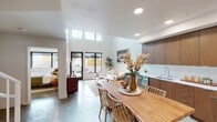 This contemporary two-floor residence showcases a thoughtfully designed open-concept layout that seamlessly connects the living, dining, and kitchen areas. The main living space commands attention with soaring double-height ceilings and clerestory windows that flood the interior with natural light, complemented by polished concrete floors that run throughout. The kitchen is outfitted with rich warm-toned wood cabinetry extending to the ceiling, white countertops, and a full suite of stainless steel appliances, including a range, microwave, dishwasher, and refrigerator. A live-edge wood dining table anchors the adjacent dining area, reinforcing the space's organic modern aesthetic. The living area offers views to an outdoor patio through large black-framed windows and glass doors. The bedrooms are well-proportioned, each featuring oversized windows that bring in ample daylight and street views. Bathrooms include a full tub-shower combination bath finished in crisp white, and a double-vanity bath with wood cabinetry and a wide mirror. The upper floor, accessible via a staircase, provides additional living or sleeping quarters. Exposed concrete ceilings in select areas add an industrial character that balances the warmth of the wood finishes throughout.