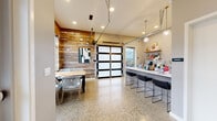 Located at 8445 Pacific Ave in Tacoma, WA, this single-floor community amenity space blends modern design with warm, natural materials throughout. The centerpiece is a dramatic great room with soaring vaulted ceilings clad in reclaimed wood planking, floor-to-ceiling windows that flood the interior with natural light, and polished terrazzo-style concrete floors that run seamlessly throughout. A statement fireplace wall, also finished in reclaimed wood, anchors the lounge area, which is furnished with comfortable seating and accented by an open outdoor view. Adjacent to the lounge, a dedicated game area features a full-sized ping pong table beneath industrial-style pendant lighting, creating a lively recreational zone. A sleek bar counter with black barstool seating sits beneath globe pendant lights, complemented by open shelving and a refreshment station. The kitchenette is finished with warm wood cabinetry, white quartz countertops, a herringbone tile backsplash, stainless steel appliances, and a deep utility sink. A large garage-style door with frosted glass panels opens the space to the outdoors, enhancing versatility and airflow. The restrooms are thoughtfully designed with decorative patterned tile flooring, full-height gray wall tile, matte black fixtures, and ADA-compliant features, maintaining the space's polished aesthetic throughout.