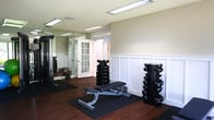 This well-appointed fitness center in Virginia Beach, VA is thoughtfully organized into distinct workout zones, offering a comprehensive exercise experience on a single floor. The strength training area showcases rich hardwood-style flooring, elegant wainscoting with classic panel molding along the walls, and an array of free weights including a full dumbbell rack, adjustable weight benches, and a cable machine — providing everything needed for a complete resistance workout. Large windows in this section flood the space with natural light and offer views of the surrounding greenery outside. The cardio zone features carpeted flooring and is equipped with multiple treadmills, elliptical trainers, and upright and recumbent stationary bikes. Recessed lighting and a beadboard-style ceiling add a polished, clean aesthetic to the cardio area. Floor-to-ceiling mirrors are strategically placed to enhance the sense of space and allow users to monitor their form. A dedicated area with additional cable and resistance machines rounds out the facility. The overall ambiance is bright, clean, and functional, with a neutral color palette of warm beige tones and white trim that creates an inviting, energizing atmosphere suited for a wide range of fitness routines.