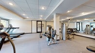 This fitness center is thoughtfully designed to support a wide range of workout routines within a clean, modern environment. The space is organized into distinct functional zones, each tailored to a specific training focus. The main strength and cardio area features a selection of Hoist selectorized machines, a full dumbbell rack with a wide range of weights, and a flat workout bench, all set against floor-to-ceiling mirrors that expand the sense of space and allow for proper form monitoring. Recessed lighting throughout the drop-tile ceiling provides consistent, well-distributed illumination across the facility. A dedicated cardio zone houses multiple elliptical trainers and a row of treadmills positioned in front of large windows that bring in natural light and offer views of the surrounding outdoor greenery. A separate studio-style room features smooth flooring, full wall mirrors, a wall-mounted television, and a rack of medicine balls — ideal for stretching, yoga, or group fitness sessions. An additional utility area provides storage and access to secondary mechanical spaces. The entry corridor is finished with neutral carpet, white walls, and a glass-paneled door with dark framing, contributing to the facility's polished and professional aesthetic throughout.