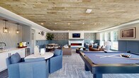The Lodges of Huntersville Gaming Center is a thoughtfully designed communal lounge space located in Huntersville, NC. The single-floor layout offers a generous, open feel that effortlessly accommodates a variety of social activities and relaxed gatherings.

The space is anchored by a centrally placed pool table, serving as the focal point of the room and setting an inviting recreational tone. A striking wood-plank ceiling adds warmth and a lodge-inspired character, complementing the slate tile flooring and layered area rugs that define each seating zone.

A standout feature is the sleek, marble-surround electric fireplace fitted with a wall-mounted television above it, framed by bold botanical-patterned wallpaper that adds a sophisticated, decorative accent. Comfortable tufted leather sofas and upholstered armchairs are arranged throughout, creating intimate conversation areas ideal for lounging.

Abundant natural light pours in through large windows and glass doors that line the perimeter, offering views of the surrounding outdoor greenery. The blue-grey wainscoting lends a refined coastal aesthetic that ties the design together cohesively.

Additional seating nooks with small round tables provide quieter spaces for conversation or casual dining. Lush indoor plants are thoughtfully placed throughout, enhancing the fresh, welcoming ambiance of this well-appointed community gathering space.