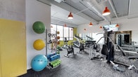 This fitness center offers a thoughtfully organized workout space designed to accommodate a wide range of exercise needs. The room features an open, single-level layout with an exposed ceiling finished in white, through which industrial-style ductwork and structural elements are visible, adding a modern, urban aesthetic. Bold red pendant lights hang throughout the space, providing focused illumination while serving as a distinctive design accent.

Large windows along one wall allow generous natural light to fill the room, offering views of the surrounding cityscape and creating an energizing atmosphere. A vibrant yellow-green accent wall adds a lively pop of color that complements the otherwise neutral white and gray palette.

The flooring consists of durable gray carpet tiles suited for high-traffic fitness use. Equipment is well-distributed throughout the space and includes stationary bikes, treadmills, elliptical machines, and a cable resistance machine. A dedicated functional training station holds an array of stability balls in multiple sizes, medicine balls, resistance bands, and rolled exercise mats. A mirrored wall section enhances the sense of openness. The overall environment is bright, modern, and practical.