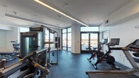 Located at 165 Huguenot St in New Rochelle, NY, this well-appointed fitness center delivers a complete workout environment within a thoughtfully designed, single-floor layout. The space is divided into distinct functional zones, seamlessly connected to create a fluid and practical exercise experience.

The cardio area is bathed in abundant natural light, courtesy of expansive floor-to-ceiling windows and glass-panel doors that open to an outdoor terrace, offering sweeping panoramic views of the surrounding New Rochelle skyline. This section is equipped with Matrix-brand treadmills, stationary cycling bikes, and additional cardio machines, alongside a wall-mounted flat-screen television. Durable rubberized blue flooring provides both comfort and safety underfoot.

The strength training zone transitions to a darker rubberized floor and features a comprehensive collection of cable machines, multi-function cable stations, and a full dumbbell rack displayed against a large wall mirror that enhances the sense of depth. Adjustable weight benches and suspension training equipment round out the free weights area. A dedicated wall-mounted storage rack holds kettlebells and medicine balls for functional training.

Recessed linear LED lighting runs along the ceilings throughout, creating a clean, modern ambiance. The crisp white walls and uncluttered layout contribute to a bright, professional atmosphere well-suited for focused training sessions.