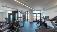 Located at 165 Huguenot St in New Rochelle, NY, this well-appointed fitness center delivers a complete workout environment within a thoughtfully designed, single-floor layout. The space is divided into distinct functional zones, seamlessly connected to create a fluid and practical exercise experience.

The cardio area is bathed in abundant natural light, courtesy of expansive floor-to-ceiling windows and glass-panel doors that open to an outdoor terrace, offering sweeping panoramic views of the surrounding New Rochelle skyline. This section is equipped with Matrix-brand treadmills, stationary cycling bikes, and additional cardio machines, alongside a wall-mounted flat-screen television. Durable rubberized blue flooring provides both comfort and safety underfoot.

The strength training zone transitions to a darker rubberized floor and features a comprehensive collection of cable machines, multi-function cable stations, and a full dumbbell rack displayed against a large wall mirror that enhances the sense of depth. Adjustable weight benches and suspension training equipment round out the free weights area. A dedicated wall-mounted storage rack holds kettlebells and medicine balls for functional training.

Recessed linear LED lighting runs along the ceilings throughout, creating a clean, modern ambiance. The crisp white walls and uncluttered layout contribute to a bright, professional atmosphere well-suited for focused training sessions.