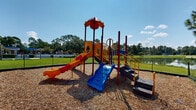 Located at 1718 Northside Dr in Valdosta, Georgia, this outdoor playground is a well-equipped recreational space designed to provide an engaging environment for children. The centerpiece of the area consists of multiple colorful, multi-level play structures featuring an array of slides in bold primary colors — including blue, red, yellow, and orange — catering to a range of ages and activity levels. The structures are topped with decorative canopy roofs and connected by platforms, railings, and climbing elements that encourage active play and physical development.

The playground surface is covered with a generous layer of wood chip mulch, providing a safe and cushioned ground cover throughout the play area. The entire space is enclosed by a chain-link fence, offering a secure environment. Accessible entry steps and ramps are integrated into the structures, enhancing usability.

Surrounding the playground is well-maintained green lawn, and the backdrop features a tranquil pond visible beyond the fence line, framed by mature trees that provide natural shade and a pleasant outdoor atmosphere. Under the open Georgia sky, the space enjoys abundant natural light, making it an inviting and lively destination for outdoor recreation in the Valdosta community.