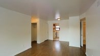This single-floor space offers a straightforward and functional layout designed for comfortable everyday living. The main living area is finished with rich, dark hardwood flooring that extends throughout, providing warmth and a classic aesthetic. White walls and a flush ceiling light keep the space feeling clean and well-lit. A notable feature of the living area is the open walk-in closet, offering generous storage space conveniently accessible from the main room. An open doorway connects the living area to a separate kitchen, which benefits from natural light streaming through a window. The kitchen is equipped with a white stove and cabinetry, providing a practical cooking environment. The overall layout is efficient and uncluttered, making good use of the available space. The neutral color palette throughout — from the white walls to the light ceiling — creates a cohesive and versatile backdrop that can be easily personalized. This space is well-suited for those seeking a functional, low-maintenance living environment with essential amenities thoughtfully arranged within a compact footprint.
