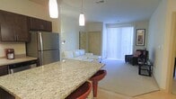This single-floor residence showcases a thoughtfully designed open-concept layout that seamlessly connects the kitchen and living areas. The kitchen is anchored by a generous granite-topped island with warm-toned bar seating, ideal for casual dining or entertaining. Stainless steel appliances, including a full-sized refrigerator, complement the sleek dark cabinetry overhead. Pendant lighting hangs above the island, adding both functional illumination and a stylish focal point to the space.

The living area flows naturally from the kitchen, offering a comfortable and welcoming atmosphere with plush carpeted flooring. A white sectional sofa serves as the centerpiece of the lounge space, accompanied by additional seating nearby. Decorative wall art adds a touch of personality to the neutral, light-toned walls.

Natural light floods the interior through a large sliding glass door dressed with vertical blinds, creating a bright and open ambiance throughout the day. The overall design balances modern finishes with a clean, neutral palette, resulting in a space that feels both contemporary and inviting. The single-floor layout ensures easy accessibility and a cohesive flow from one area to the next.