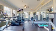 The Columns Fitness Center at 4330 Point Cypress Blvd in Wesley Chapel, Florida, is a thoughtfully designed, single-floor community gym that combines functionality with a bright, welcoming atmosphere. The space features soft sage-green walls accented by crisp white crown molding and wainscoting, lending a clean, polished aesthetic throughout. Large, multi-pane windows line the walls on multiple sides, flooding the interior with abundant natural light and offering pleasant views of the surrounding landscape and community pool area. A ceiling fan with a classic fixture provides comfortable air circulation. Full-length wall mirrors on two sides amplify the sense of space and allow for proper form checks during workouts. The fitness center is equipped with a comprehensive selection of TRUE-brand cardio machines, including multiple treadmills and elliptical trainers, as well as a multi-station cable strength machine, a leg extension/curl bench, a stationary bike, and free weights. Step platforms and additional accessories support group-style or individual training routines. Wall-mounted flat-screen televisions provide entertainment during workouts. The durable rubber flooring throughout ensures safety and comfort. A dedicated entry door with an exterior light fixture provides convenient access to the outdoors. Sanitation stations and informational signage round out the practical amenities of this well-maintained fitness facility.