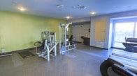 This fitness room offers a functional and well-equipped workout space designed to support a range of strength training exercises. The room features a prominent multi-station cable and weight machine as its centerpiece, complemented by additional selectorized strength equipment positioned nearby, providing users with versatile upper and lower body training options. The flooring is finished in a durable speckled composite material, which is both practical and easy to maintain, with designated mat areas offering cushioned zones for floor-based exercises. The walls are painted in a fresh lime green accent, adding a vibrant, energizing atmosphere to the space, while the remaining surfaces are finished in neutral grey tones. Recessed and track lighting mounted on the ceiling provides even illumination throughout the room. Toward the rear of the space, a small utility alcove features dark cabinetry, a countertop with a sink or hand-washing station, and wall-mounted dispensers, adding a convenient hygiene area for users. A yellow utility door on the far wall provides secondary access to the space. A wall-mounted vent near the baseboard supports climate control, contributing to a comfortable environment during workouts.