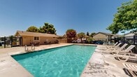 Located at 1180 Matmor Rd in Woodland, California, this outdoor community pool area offers a well-appointed recreational space designed for relaxation and leisure. The centerpiece of the area is a generously sized rectangular swimming pool with clear, blue water, accessible via stainless steel entry rails on multiple sides. The pool is surrounded by a spacious concrete sun deck that provides ample room for movement and lounging. A row of white lounge chairs lines one side of the pool, offering dedicated spaces for sunbathing under the California sun. On the opposite side of the deck, a patio seating area with tables and chairs provides a comfortable spot for outdoor dining or socializing. A single-story pool house or utility building is situated adjacent to the deck, likely housing restrooms or storage facilities. The entire pool area is enclosed by a dark metal security fence, ensuring safety and privacy. Mature trees, including a vibrant flowering crepe myrtle, are positioned around the perimeter, providing natural shade and adding color to the space. The surrounding residential buildings are visible beyond the fence, giving context to this shared community amenity. The area benefits from an open-sky setting, maximizing natural light throughout the day.