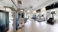 Located at 3100 Van Buren Blvd in Riverside, CA, this well-appointed fitness center delivers a comprehensive workout environment within a bright, single-floor layout. The space features light hardwood-style flooring throughout, crisp white walls, and a tray ceiling with a large ceiling fan that promotes airflow and comfort during exercise sessions. Abundant natural light streams in through multiple large windows and glass doors, offering views of the surrounding outdoor greenery and creating an open, inviting atmosphere. Wall-mounted flat-screen TVs are positioned for easy viewing from the cardio equipment area. The facility is equipped with a full lineup of Matrix-branded cardio machines, including treadmills, ellipticals, a stair climber, and a stationary bike. Strength training is well-represented with a Matrix multi-function cable machine, a leg press, an adjustable weight bench, and a dumbbell rack. A dedicated Training Zone area offers TRX suspension trainers and functional fitness accessories mounted to a wall-integrated rack. A striking green abstract mural anchors one accent wall, adding a bold visual element to the space. Full-length mirrors along the walls enhance the sense of space and support proper form during workouts.