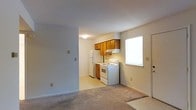 This single-floor residential unit offers a practical and straightforward layout designed for comfortable everyday living. The living area features neutral-toned carpeting that creates a warm, understated foundation for the space, and the open layout connects seamlessly to the kitchen, promoting a sense of openness and ease of movement.

The kitchen is efficiently arranged and includes warm-toned wooden upper cabinetry that provides ample storage. White appliances — including a refrigerator, electric range with a ventilation hood, and additional under-counter space — are neatly positioned along the kitchen wall. A window adjacent to the range allows natural light to fill the kitchen area, brightening the workspace. The kitchen floor transitions to a light-colored vinyl or tile surface, offering a clean contrast to the carpeted living space.

The walls throughout are finished in a soft, neutral tone that contributes to a calm and airy ambiance. Light switches and electrical outlets are thoughtfully placed, and a flush-mount ceiling light provides functional overhead illumination. A door near the kitchen suggests convenient access to an exterior or adjacent space. Overall, this unit presents a clean, functional environment well-suited for day-to-day living.