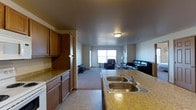 This single-floor space showcases an open-concept layout that connects the kitchen and living area in one cohesive flow. The kitchen is outfitted with warm medium-toned wood cabinetry offering generous storage, complemented by speckled granite-style countertops that extend to a peninsula with a double stainless steel sink and a chrome faucet. Light tile flooring anchors the kitchen zone, providing a practical and easy-to-maintain surface. The transition into the living area is seamless, where plush carpeting creates a comfortable, inviting atmosphere underfoot. A window on the far wall allows natural light to fill the living space, brightening the neutral-toned walls and creating an open, airy feel. The living area provides ample room for seating arrangements, making it well-suited for both relaxing and entertaining. Flush-mount ceiling lighting serves both zones, ensuring consistent illumination throughout. The overall design balances functionality with comfort, featuring a practical layout that maximizes the use of the available space across both the culinary and leisure areas.