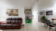 This thoughtfully designed single-floor residence offers an efficient and functional layout across a combined living and dining area, one bedroom, a kitchen, and a full bathroom. The open living and dining space is highlighted by light hardwood-style flooring and a striking faux stone accent wall that adds texture and character to the room. The living area provides ample space for seating and entertaining. The adjacent dining area features wainscoting, wall-mounted lighting, and continued stone wall accents that create a cohesive aesthetic throughout the common areas. The galley-style kitchen is equipped with white cabinetry, a light countertop, and a bold blue mosaic tile backsplash that adds a vibrant design element. Open shelving provides additional storage and display space. The bedroom is carpeted for warmth and comfort, featuring a ceiling fan, a sliding-door closet, and multiple windows dressed with curtains that allow natural light to filter in. The bathroom features painted concrete block walls, built-in cabinetry for storage, and light flooring that keeps the space bright. Throughout the residence, the interplay of clean white walls, natural-toned flooring, and rustic stone accents creates a warm and inviting atmosphere.